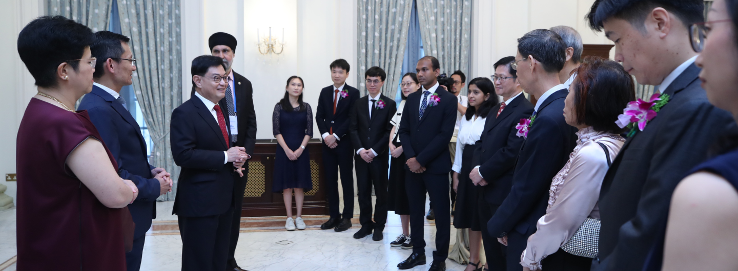 Group of people in formal wear, some with flower corsages, standing in a light-filled room.